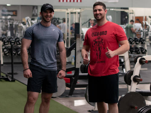 two men are standing and smiling in a gym. one is wearing a blue shirt and black shorts, while the other is in a red shirt and black shorts, giving a thumbs up. gym equipment is visible behind them.